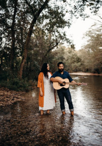Two people stand in a river. One wears an orange and white dress. The other is dressed in black and holds a guitar.
