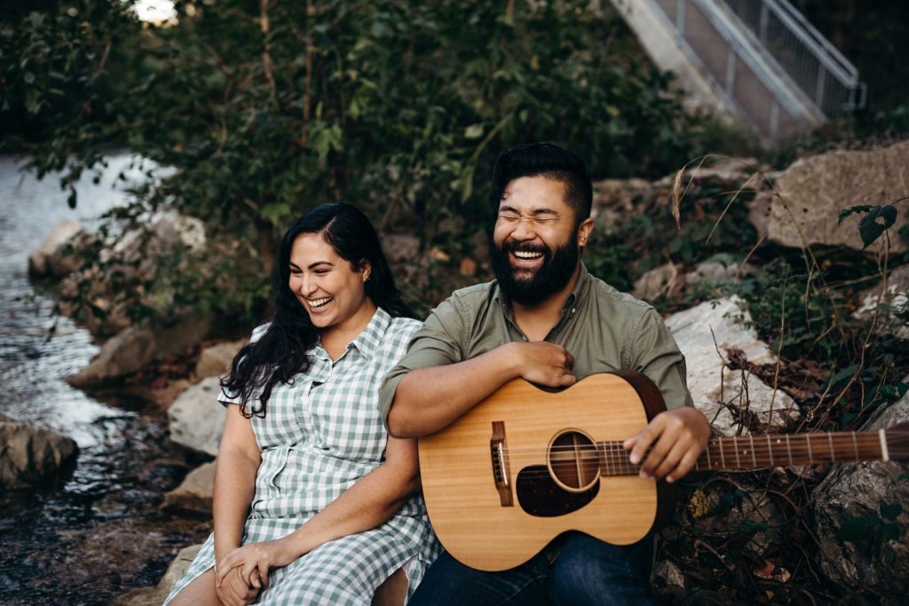 Two people sit together by a river. One wears a white dress, the other has a green shirt and is holding a guitar.