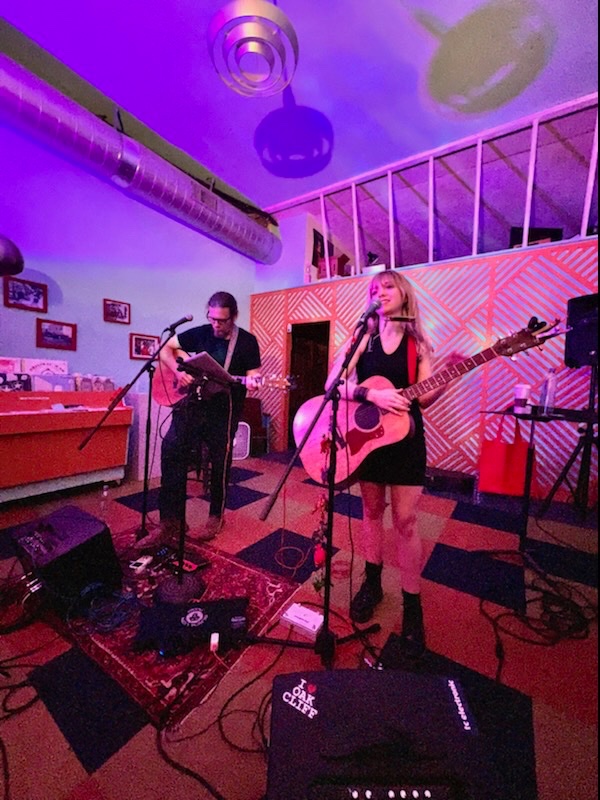 two musicians playing guitars in a pink room with purple lights.