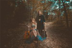 A woman with a guitar sits on a forest path next to man who is stranding and wearing a black jacket.