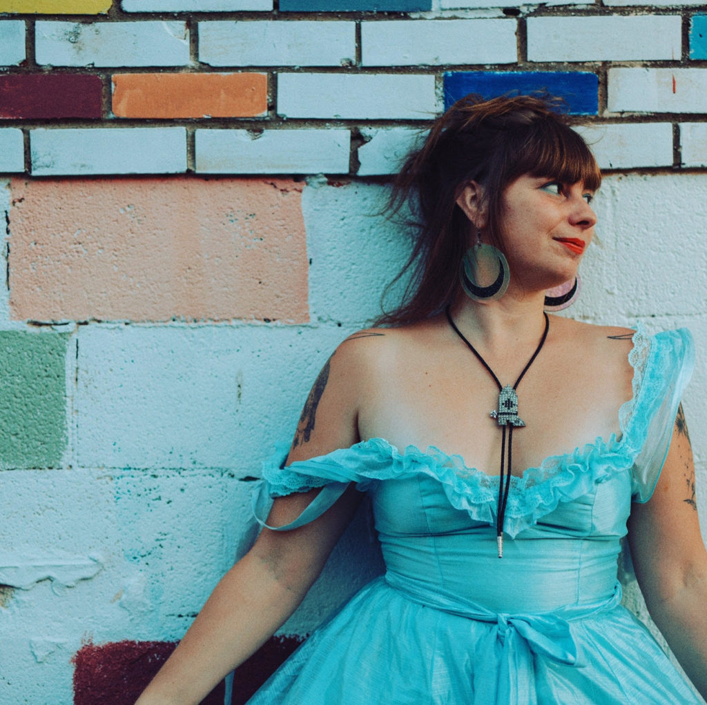 A woman in a turquoise dress leans against a painted brick wall.