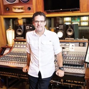 A man wearing a white shirt and glasses leans against a mixing board.