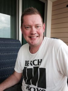 Man with short hair, wearing a white t-shirt with black design, smiles at the camera.