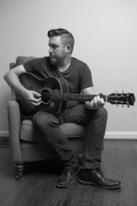 A man in a t-shirt and jeans sits in an armchair while playing guitar.