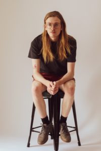 Person with long hair and glasses sitting a stool and looking at the camera.