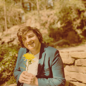 A man in a green blazer holds a large yellow flower and smiles.