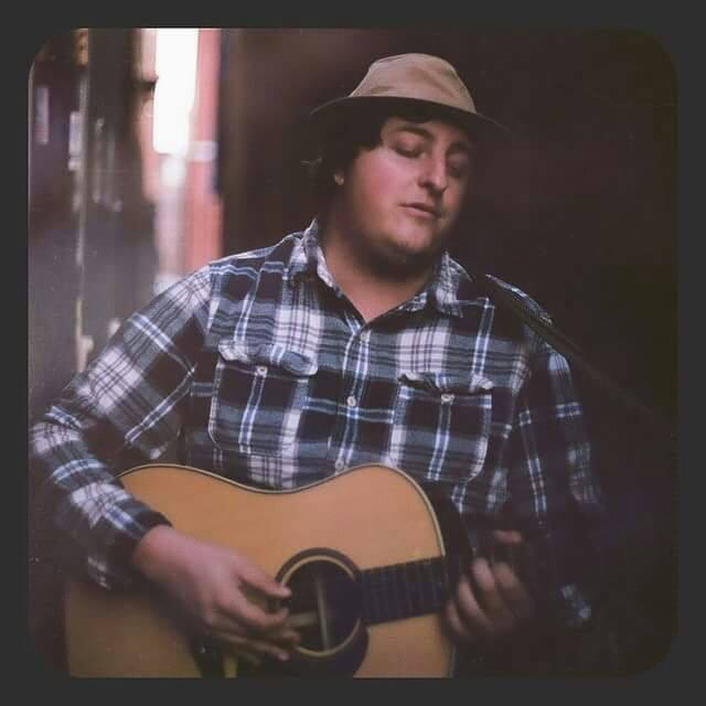 A man in a tan hat and plaid shirt plays guitar while looking away from the camera.