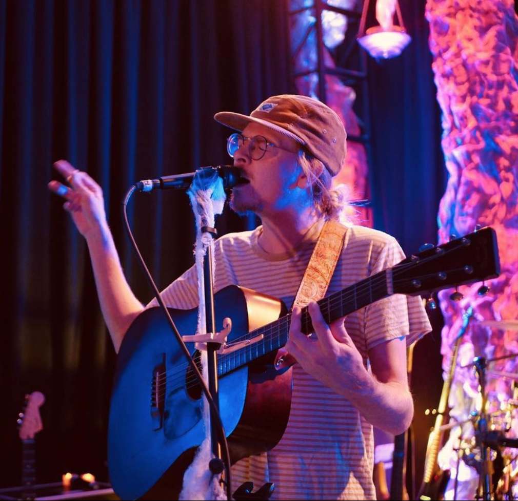 Musician with long hair and cap holding a guitar and singing into a microphone.