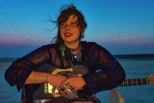 A woman in a black dress holds a guitar in front of a blue evening sky.