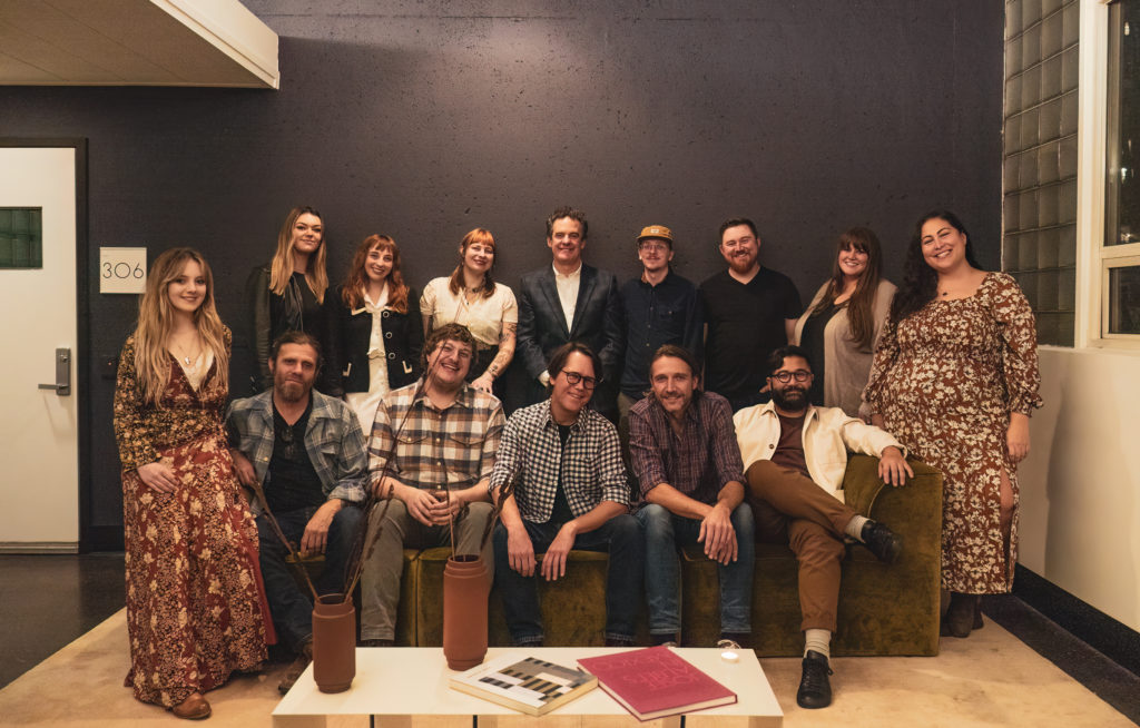 A group of 14 people who competed in the NewSong Music Competition stand behind and sit on a green couch.