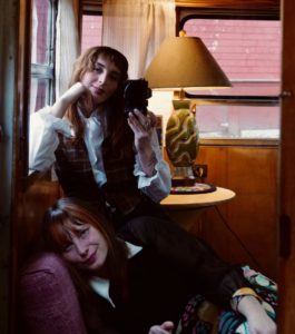 Two women, Kendall Lujan and AC Sapphire, sit on a couch in front of a yellow lamp. Kendall holds a vintage camera.