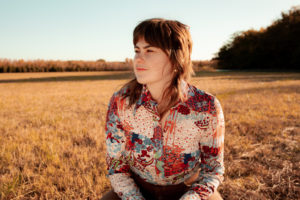 Jobi Riccio wears a colorful vintage shirt and sits in a field.