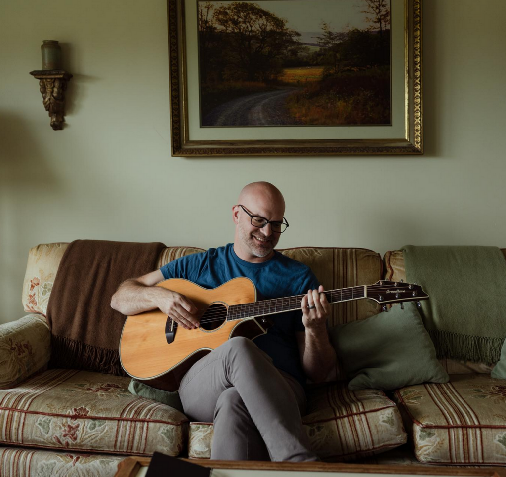 Nathan Storey sitting on a sofa holding a guitar.